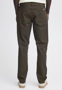 Pantalones de mezclilla marrón oscuro con un diseño de pierna recta, que cuentan con dos bolsillos traseros con un patrón de puntada en zigzag, usados con zapatos de color claro.
