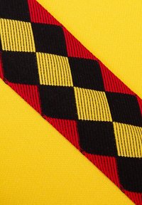 Geel stof met een diagonaal rood, zwart en geel ruitpatroon. Textuurstiksel zichtbaar langs de randen van het patroon.