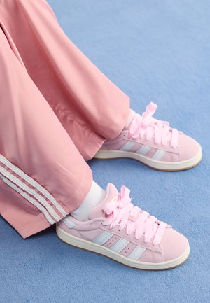 Baskets de sport roses avec des rayures bleu clair, bout rond et lacets épais roses, associées à un pantalon de survêtement rose avec des rayures blanches sur les côtés.
