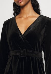 Svart sammetsinlindad klänning med V-ringning och långa ärmar, utrustad med ett brett bälte med fyrkantig spänne vid midjan. Slät textur.