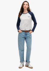 Jonge vrouw in een lichtgrijs en marineblauw shirt met lange mouwen, lichtblauwe opgerolde spijkerbroek, witte sneakers en een bruine riem, staand met handen in de zakken.