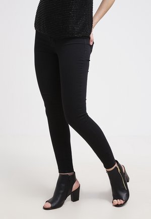 Personne portant un pantalon skinny noir et des bottines à talons ouvertes noires avec un détail de fermeture éclair dorée sur fond blanc.