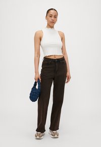Hvit ribbet crop-top med bølgete kanter, parret med brune høytlivsjeans, holder en blå vevd håndveske, og har på seg beige joggesko.