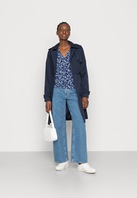 Um casaco trench navy sobre uma blusa floral, combinado com jeans de perna larga azul claro e ténis brancos. Segurando uma bolsa branca acolchoada.