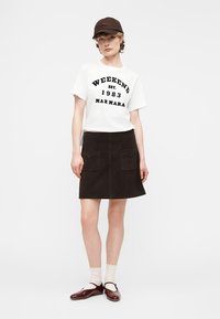 Γυναίκα που φοράει λευκό μπλουζάκι με την επιγραφή «WEEKEND EST. 1983 MAX MARA», σκουρόχρωμη φούστα με μπροστινές τσέπες, καφέ καπέλο, λευκές κάλτσες και καφέ παπούτσια Mary Jane.