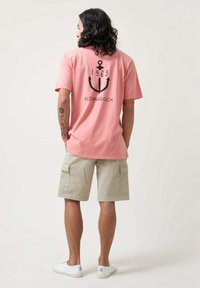 Camiseta de algodón rosa con gráfico de ancla negra y el texto "ALTONADOCK 1969" en la parte trasera, combinada con pantalones cortos de carga beige y zapatillas blancas.
