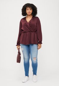 A woman with curly hair wears a burgundy top and ripped jeans, holding a maroon handbag, standing against a plain white background.