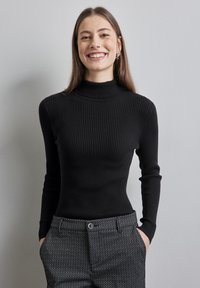 Maglione nero a costine con collo alto e maniche lunghe. Indossato con pantaloni grigi a fantasia che presentano una leggera lucentezza. Materiale in maglia leggera.