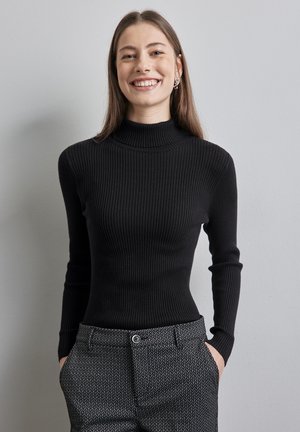 Maglione nero a costine con collo alto e maniche lunghe. Indossato con pantaloni grigi a fantasia che presentano una leggera lucentezza. Materiale in maglia leggera.