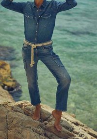 Persona descalza, vistiendo una camisa de mezclilla y jeans con un cinturón de cuerda, de pie en una orilla rocosa junto a agua clara con los brazos levantados.