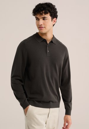 Jeune homme regardant sur le côté portant un polo à manches longues foncé et un pantalon clair sur un fond uni.