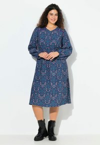 Frau mit lockigem Haar, die lächelt, trägt ein langärmliges, blau gemustertes Kleid und schwarze Schnürstiefel, steht vor einem weißen Hintergrund.