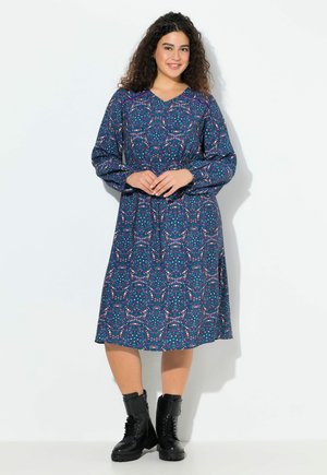 Frau mit lockigem Haar, die lächelt, trägt ein langärmliges, blau gemustertes Kleid und schwarze Schnürstiefel, steht vor einem weißen Hintergrund.