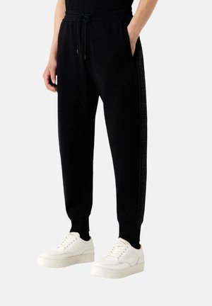 Sorte joggerbukser med snørelukning i taljen, sidelommer og tekstureret sort stribe ned ad benet, parret med hvide sneakers.