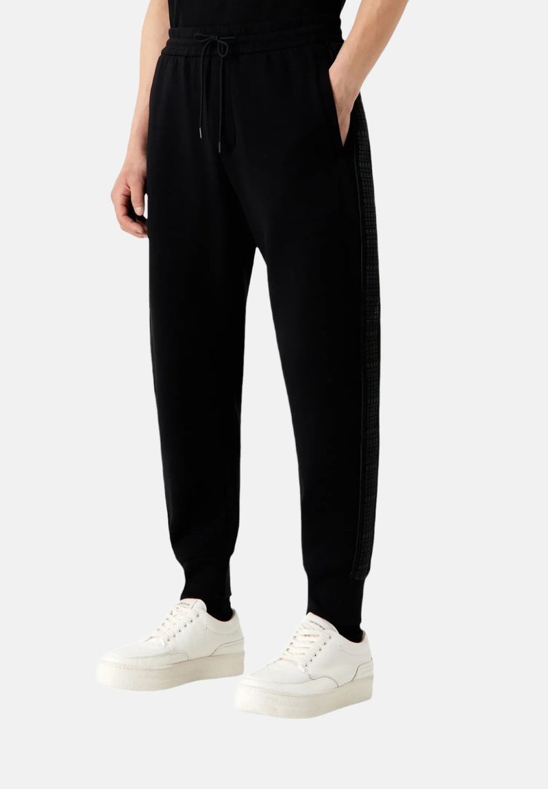 Pantaloni da jogging neri con vita regolabile, tasche laterali e una striscia nera texture lungo la gamba, abbinati a sneakers bianche.
