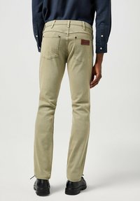 Pantalones de mezclilla beige con corte recto, bolsillos traseros y un parche de cuero en la cintura. Combinados con una camisa azul marina y zapatos negros.