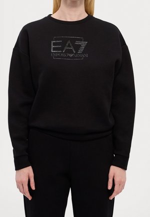 Black long-sleeve sweatshirt with "EA7 Emporio Armani" logo in sparkly silver lettering on front, worn by a person with blonde hair.