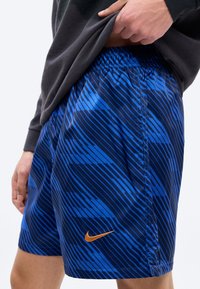 Close-up van een persoon die een donker shirt optilt om een blauwe Nike-short met diagonale strepen en een oranje swoosh-logo op de dij te laten zien.