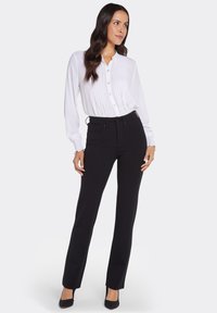 Vit plisserad blus med knappknäppning framtill, kombinerad med svarta smala byxor. Modellen bär svarta klackar; slätt tyg och skräddarsydd design.