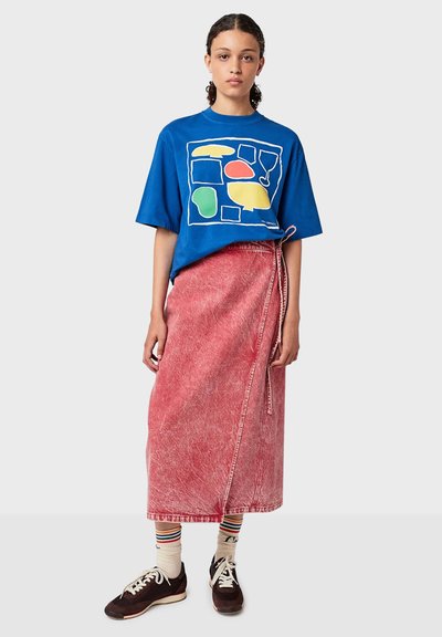 Une jeune personne se tenant debout, portant un t-shirt graphique bleu, une jupe-culotte rouge, des chaussettes rayées et des baskets noires devant un fond uni.