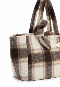 Sac fourre-tout en tissu à carreaux marron et beige, avec une poignée nouée et un petit médaillon rond en or suspendu à une chaîne.