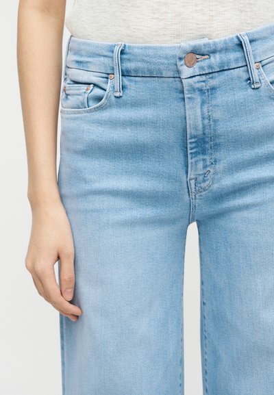 Άτομο που φοράει ανοιχτό μπλε τζιν ψηλόμεσο με ορατά κουμπιά και μπροστινές τσέπες, όρθιο σε απλό φόντο.