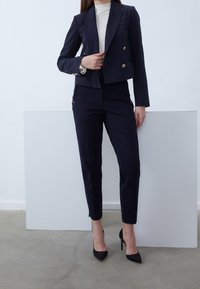 Costume bleu marine sur mesure avec une veste courte croisée et un pantalon slim, agrémenté de boutons dorés et de talons noirs pointus.