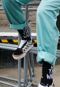 Las zapatillas Vans en blanco y negro presentan un patrón a cuadros, cordones blancos y una suela marrón clara, combinadas con calcetines negros y pantalones turquesa.