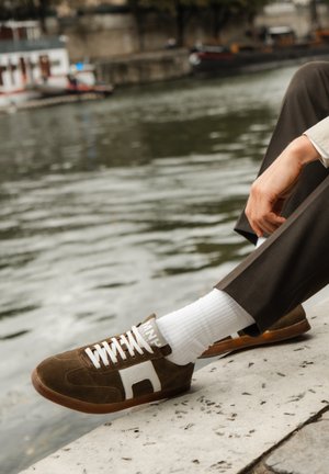 Braune Wildleder-Sneaker mit weißen Akzenten, weißen Schnürsenkeln und Gummisohle. Tragen mit weißen gerippten Socken und braunen Hosen am Ufer eines Flusses.