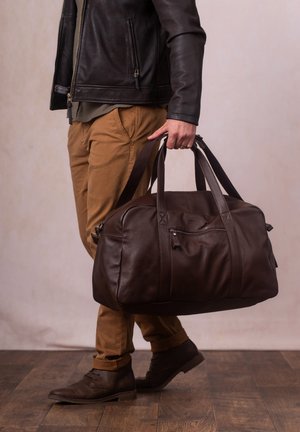 Dark brown leather duffel bag with dual handles, zippered front pocket, stitched accents, and smooth texture, carried by a person in casual attire.