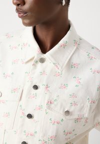 Veste en denim florale en crème avec des fleurs roses et des accents verts. Présente un col pointu, des boutons-pression et des poches frontales. Tissu texturé.
