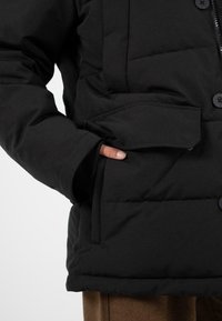 Černá péřová bunda na zip a s knoflíkovým zapínáním, s náprsní kapsou a žebrovanými manžetami, vyrobená z texturované, zateplené látky.