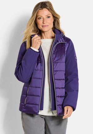 Femme portant une veste matelassée violette sur un pull blanc, tenant le col de la veste, debout devant un fond clair.