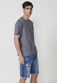 Sivi pamučni T-shirt s okruglim izrezom i prednjom vezom, uparen s plavim traper kratkim hlačama s istrošenim završetkom i preokrenutim rubovima.