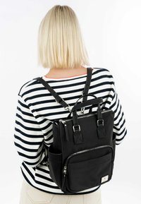 Black ribbed fabric backpack with adjustable straps, zippered pockets, and top handle, worn on the back of a person in a striped sweater.