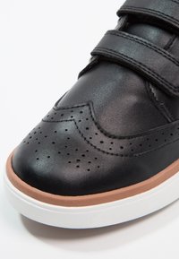 Baskets en cuir noir avec un design perforé, des straps Velcro et une semelle intermédiaire en caoutchouc brun, dotées d'une semelle extérieure en caoutchouc blanc.