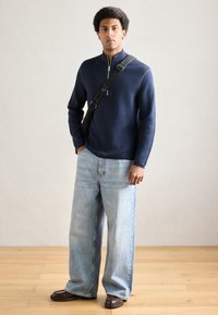 Tengerészkék cipzáras pulóver magas gallérral, világoskék bőszárú farmerrel és barna cipővel párosítva. Sima textúra és minimális részletek jellemzik.