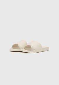 Beige Schlupfsandalen mit offener Zehenpartie. Weiche, strukturierte Riemen mit einem dezenten Muster. Flache Sohle mit einem minimalistischen Design.