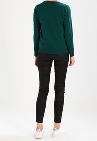 Green sweater, long sleeves, ribbed hem; paired with high-waisted black jeans, fitted design; white sneakers with decorative accents.