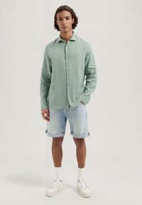 Camisa de lino verde claro con botones y mangas largas, pantalones cortos de mezclilla azul claro enrollados y zapatillas blancas con cordones visibles y acentos.