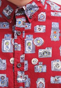 Red short-sleeve shirt featuring a camera print, with blue and yellow accents. Button-down collar and textured fabric.