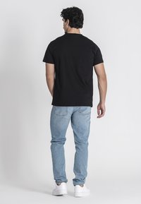 Crna majica s kratkim rukavima, pamučni materijal, jednostavan dizajn, uparen s svijetloplavim trapericama i bijelim tenisicama, prikazana sa stražnje strane.