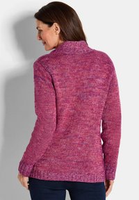 Frau, die sich von hinten zeigt, trägt einen lila-pinken Strickpullover mit langen Ärmeln und dunkler Hose vor einem schlichten Hintergrund.