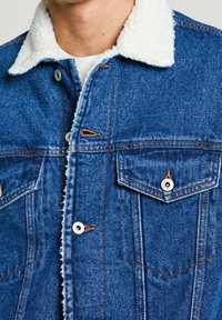 Blå denimjacka med en vit, fluffig krage. Har två bröstfickor, silverfärgade tryckknappar och orange stickning vid sömmarna.