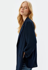 Navy blue blazer made from a lightweight fabric with a relaxed fit, featuring a notched lapel, side pockets, and a three-button closure.