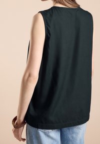 Bezrekawnik w ciemnozielonym kolorze o gładkiej fakturze, z okrągłym dekoltem i luźnym krojem, zestawiony z jasnoniebieskimi dżinsami. Przezroczysta brązowa bransoletka jako dodatek.