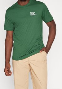 Zelené bavlněné tričko s kulatým výstřihem. Na levém prsu je bíly text "UMÍTE MLUVNOUT LACOSTE?". Kombinováno s béžovými kalhotami.