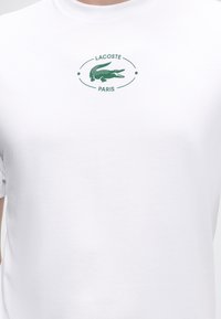 Λευκό T-shirt με πράσινο λογότυπο κροκόδειλου Lacoste στο κέντρο, συνοδευόμενο από το κείμενο "LACOSTE PARIS" πάνω και κάτω από το λογότυπο.