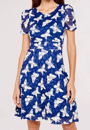 Kobieta ubrana w niebieską sukienkę o długości do kolan z krótkimi bufkami i białymi motylami na całej tkaninie.