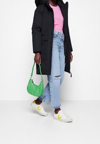 Abrigo negro con capucha, camiseta rosa, jeans claros desgastados, bolso verde y zapatillas blancas con detalles amarillos.